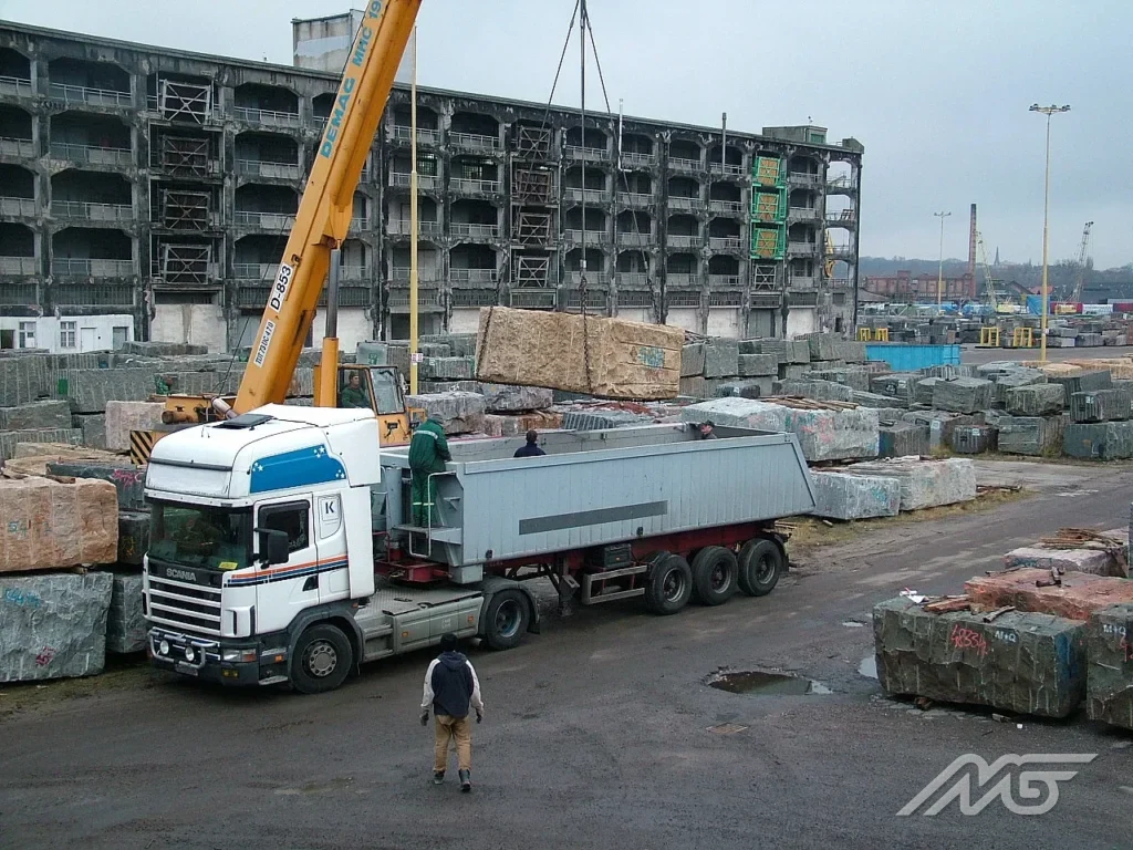Crane lowering a stone block onto a tipper semitrailer at a storage yard